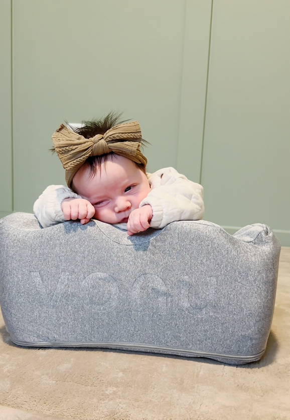 Baby with a bow headband leaning on a gray cushion.