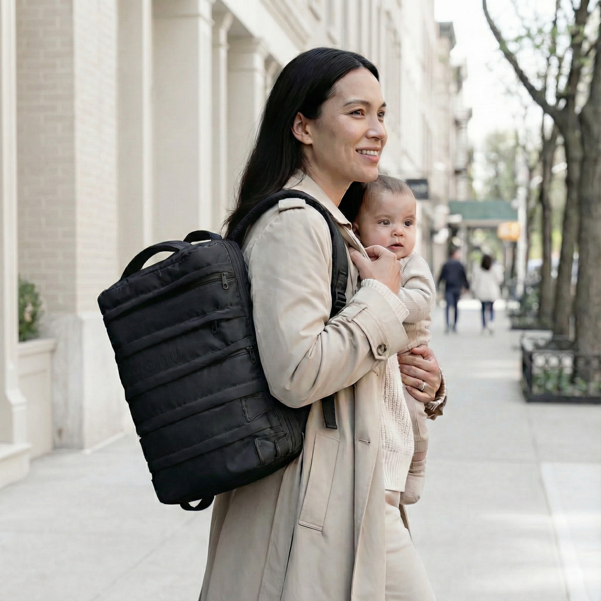 Woman carrying a baby and wearing a black backpack, walking on a city sidewalk.