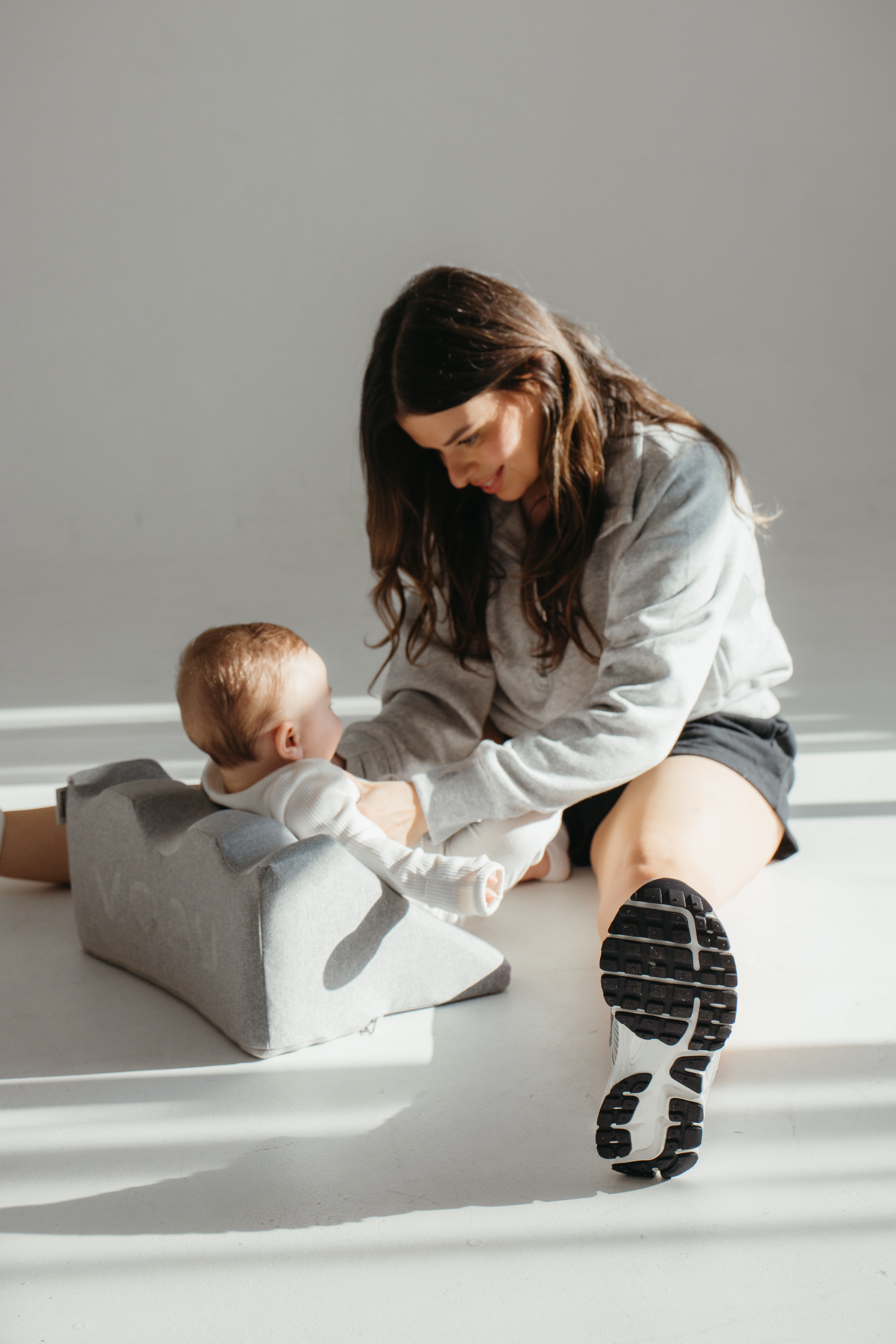 A woman sitting on the floor, playing with a baby using a grey support cushion.