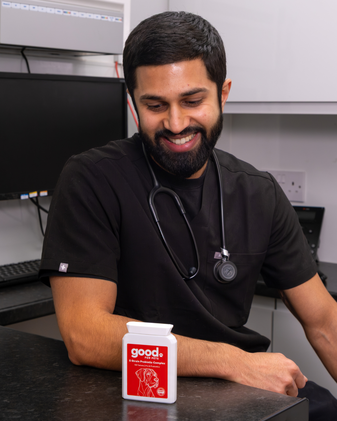 A smiling veterinarian in black scrubs looks down at a bottle of 'good for pets' supplements.