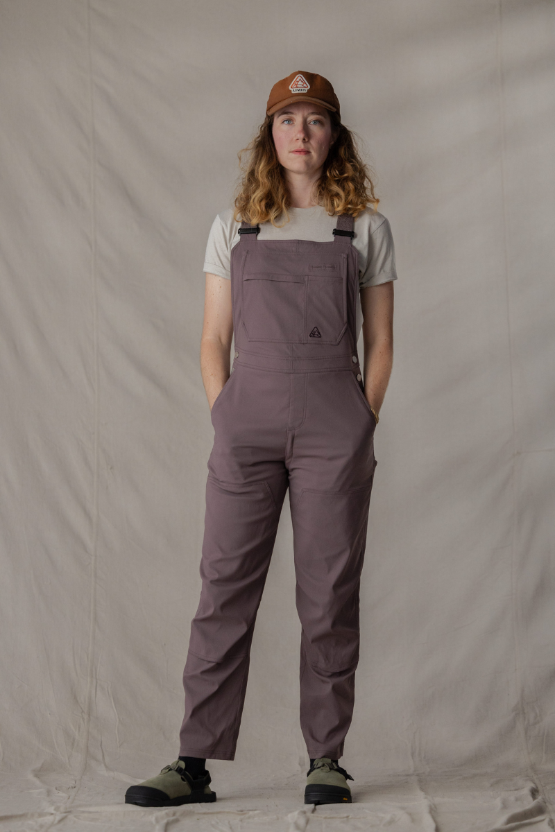 Person wearing a brown cap, beige shirt, and purple overalls against a plain background.