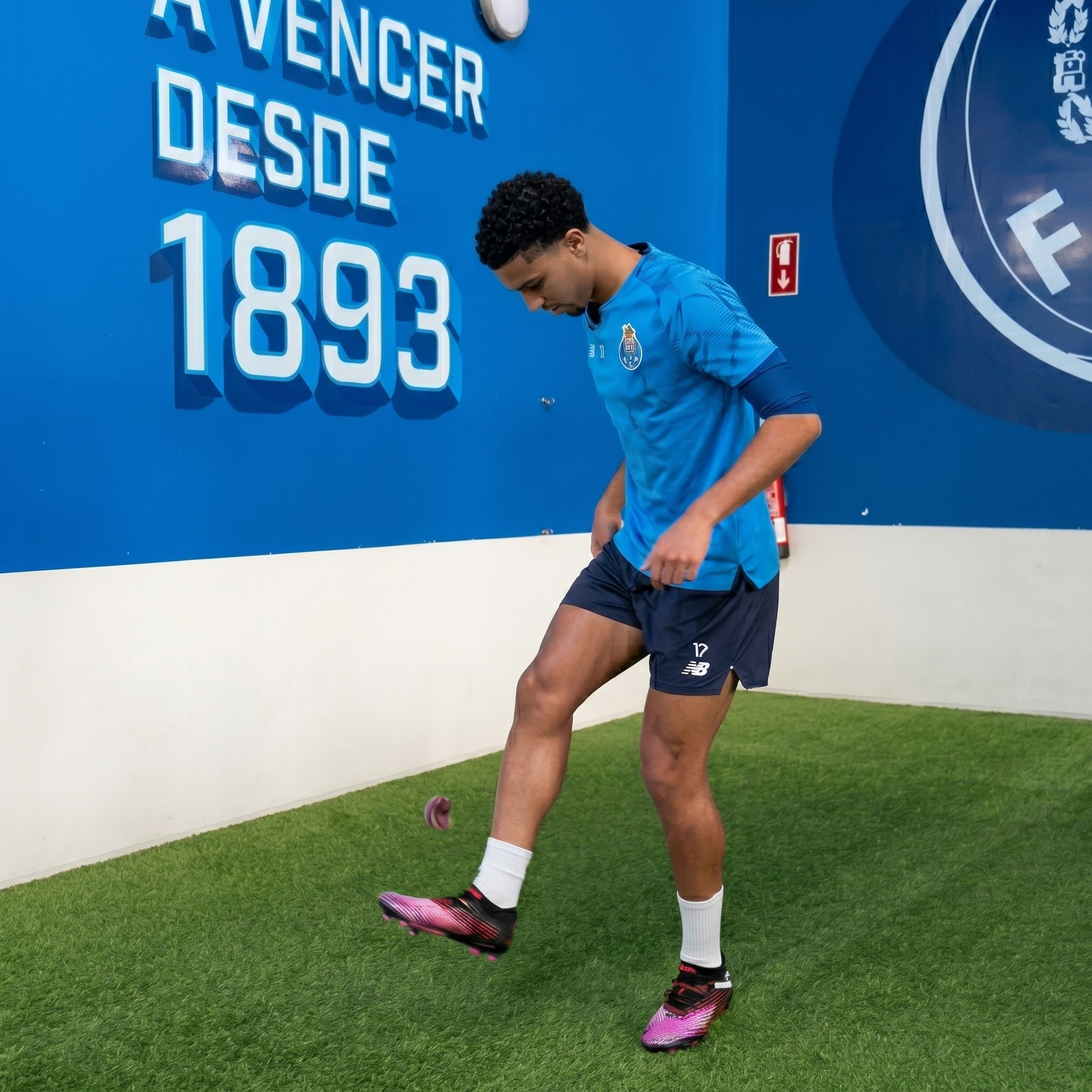 A soccer player in a blue kit juggles a small ball on an indoor artificial turf pitch.