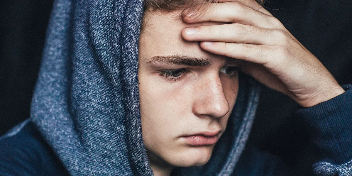 A young man in a blue hoodie rests his head on his hand, looking down thoughtfully.