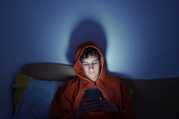 A young man in an orange hoodie looks at his phone in a dark room, his face illuminated by the screen.