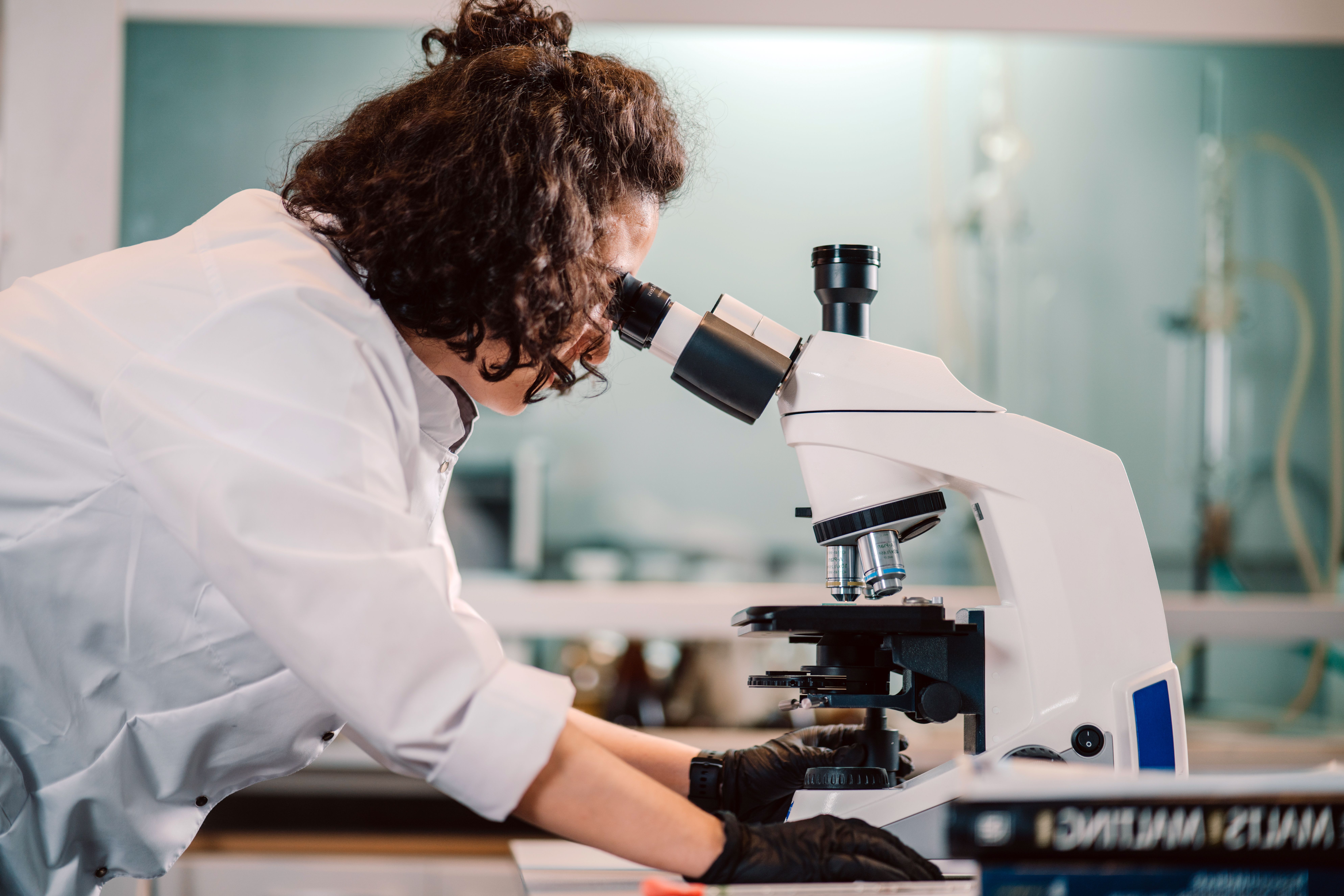A researcher in a white lab coat and black gloves looks into a microscope in a laboratory.