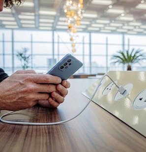 A person holds a blue smartphone while it charges at a public charging station on a wooden table.