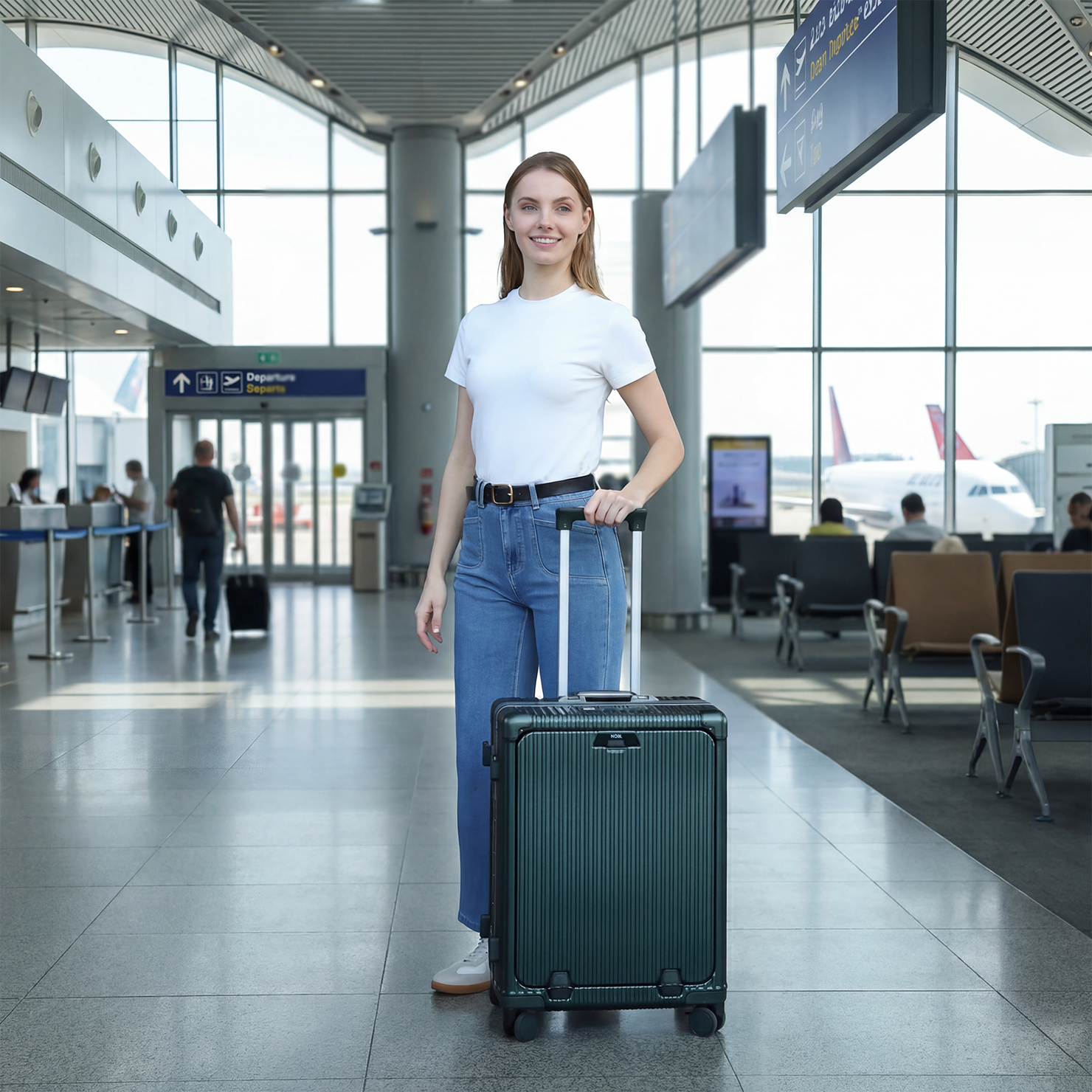 A woman in a white t-shirt and jeans stands with a dark green rolling suitcase in an airport terminal.