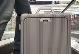 A gray, ribbed suitcase sits on a moving walkway next to a person's leg.