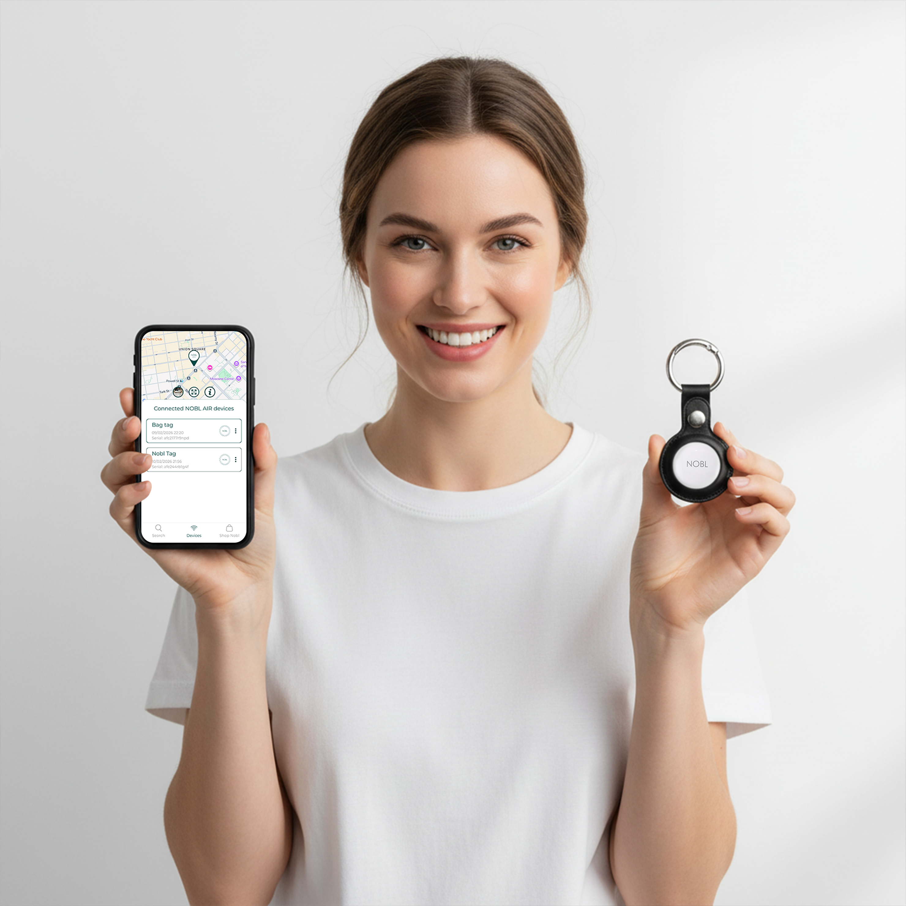 A smiling woman holds a smartphone with a tracking app and a NOBL tracking tag on a keychain.