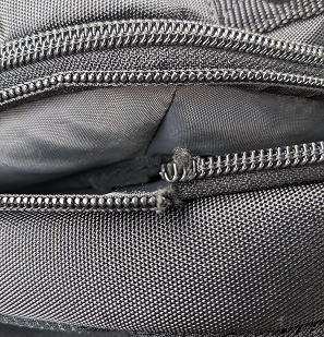 A close-up of a broken zipper on a black fabric bag, showing damaged and separated teeth.