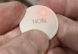 A person's fingers holding a small, white, circular device with the word NOBL and a glowing light.
