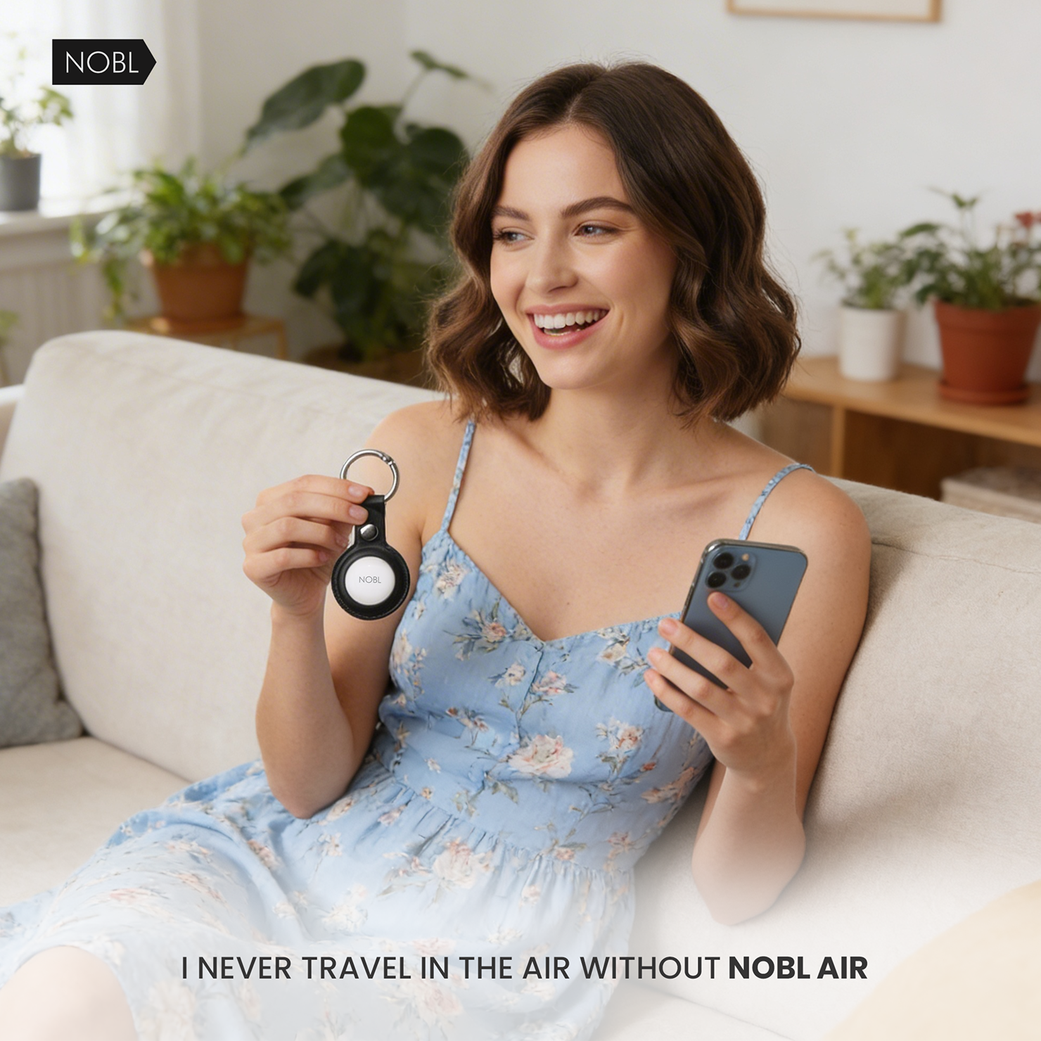 A smiling woman sits on a couch, holding a smartphone and a NOBL AirTag holder keychain.