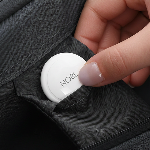 A hand placing a white, circular NOBL tracker into an inner pocket of a grey bag.