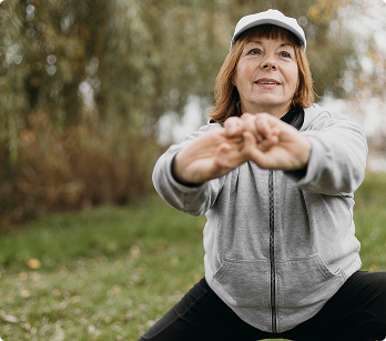 An older woman wearing a white cap and grey hoodie stretches outdoors in a park.