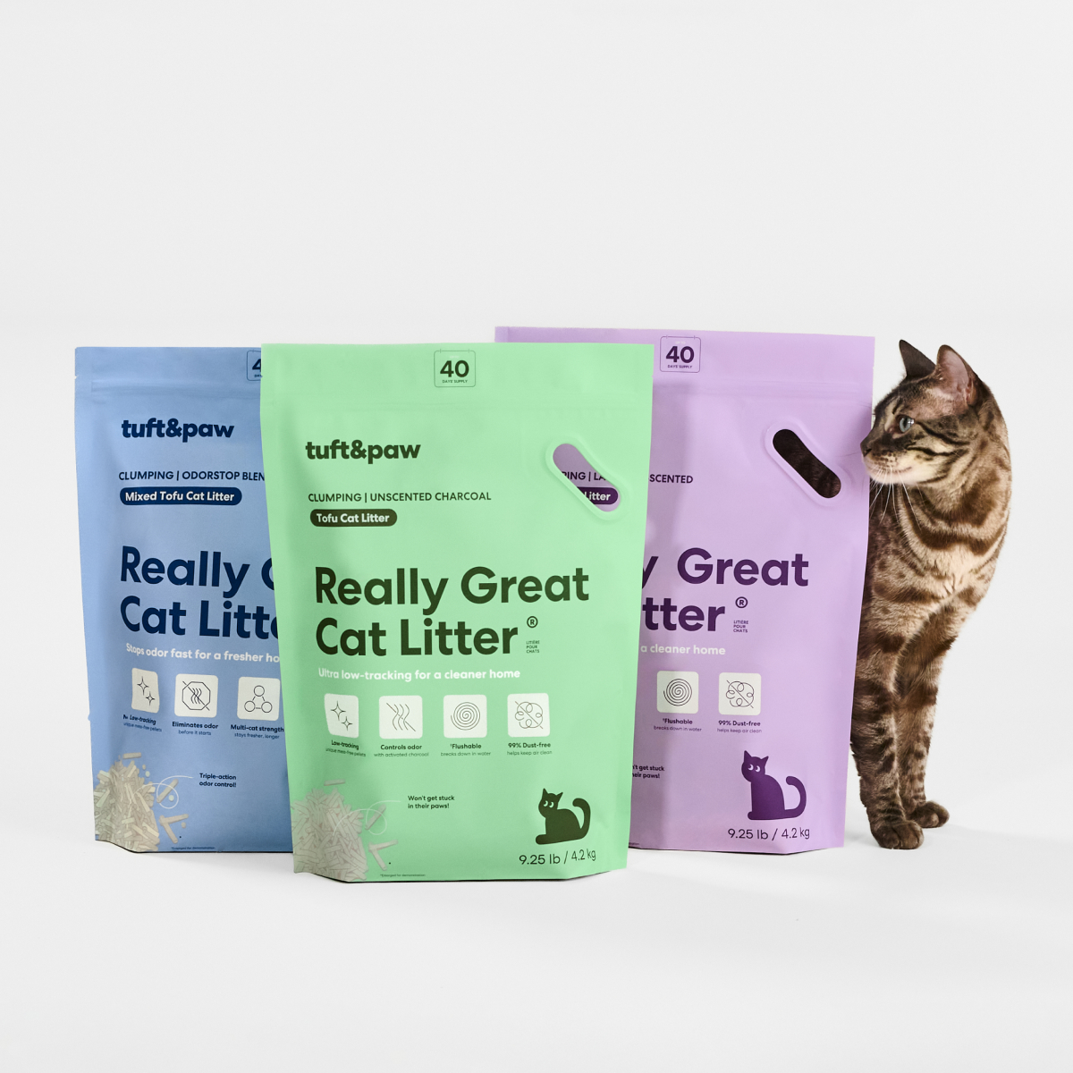 A brown tabby cat stands next to three colorful bags of tuft&paw Really Great Cat Litter.