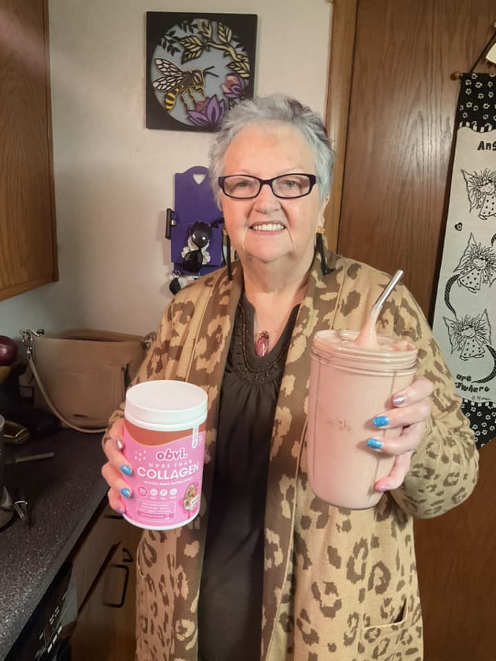 Person holding a collagen supplement container and a smoothie in a kitchen.