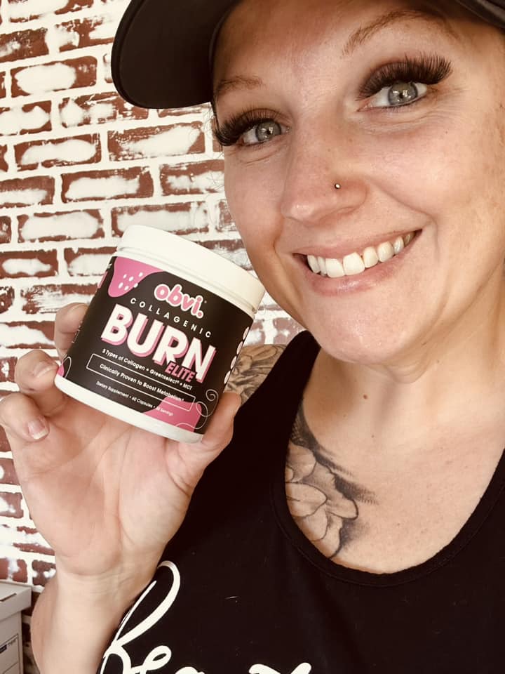 Person smiling and holding a container labeled 'Obvi Collagenic Burn Elite' against a brick wall background.