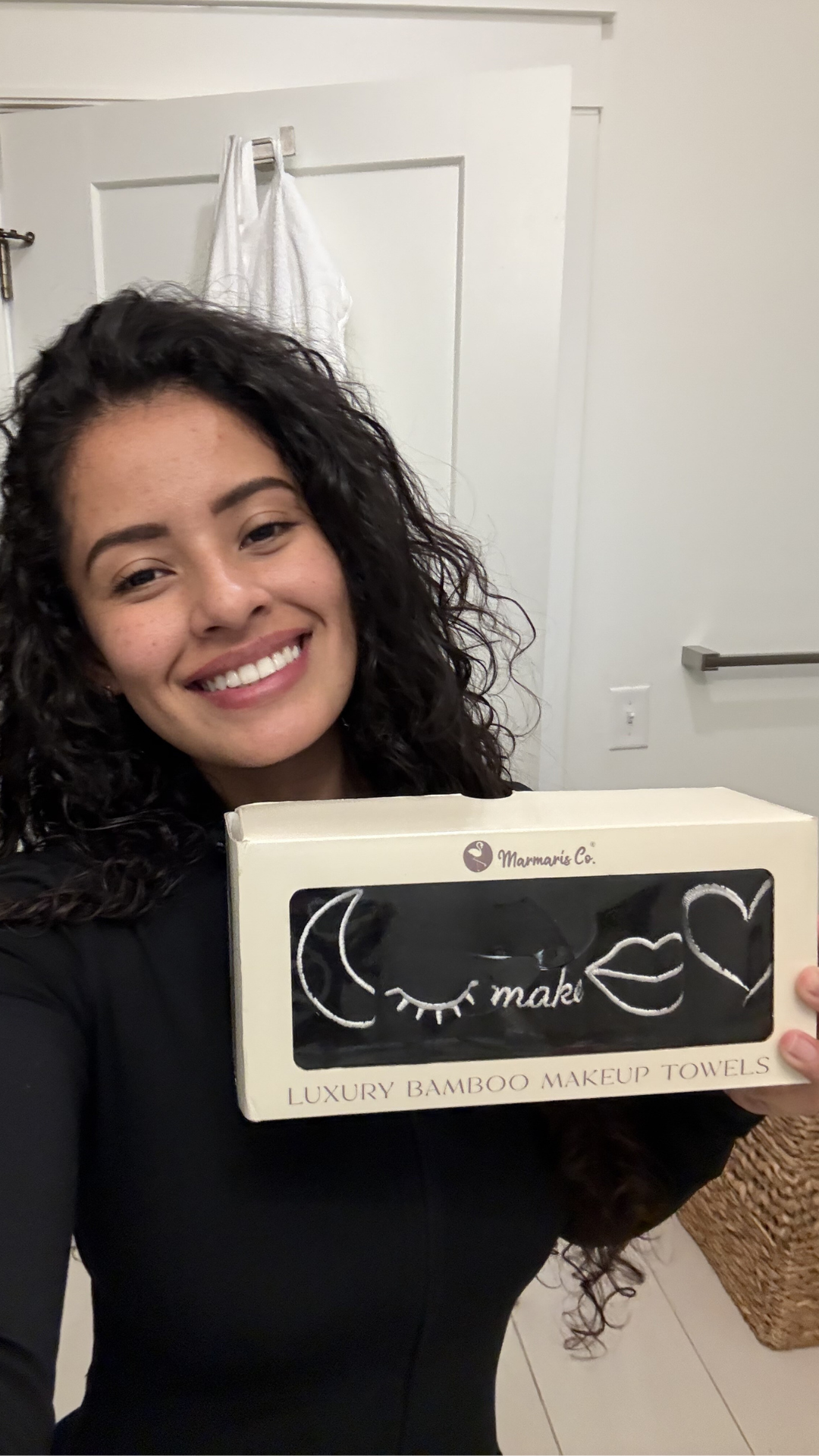 A smiling woman holds a box of Marmaris Co. Luxury Bamboo Makeup Towels in a bathroom.