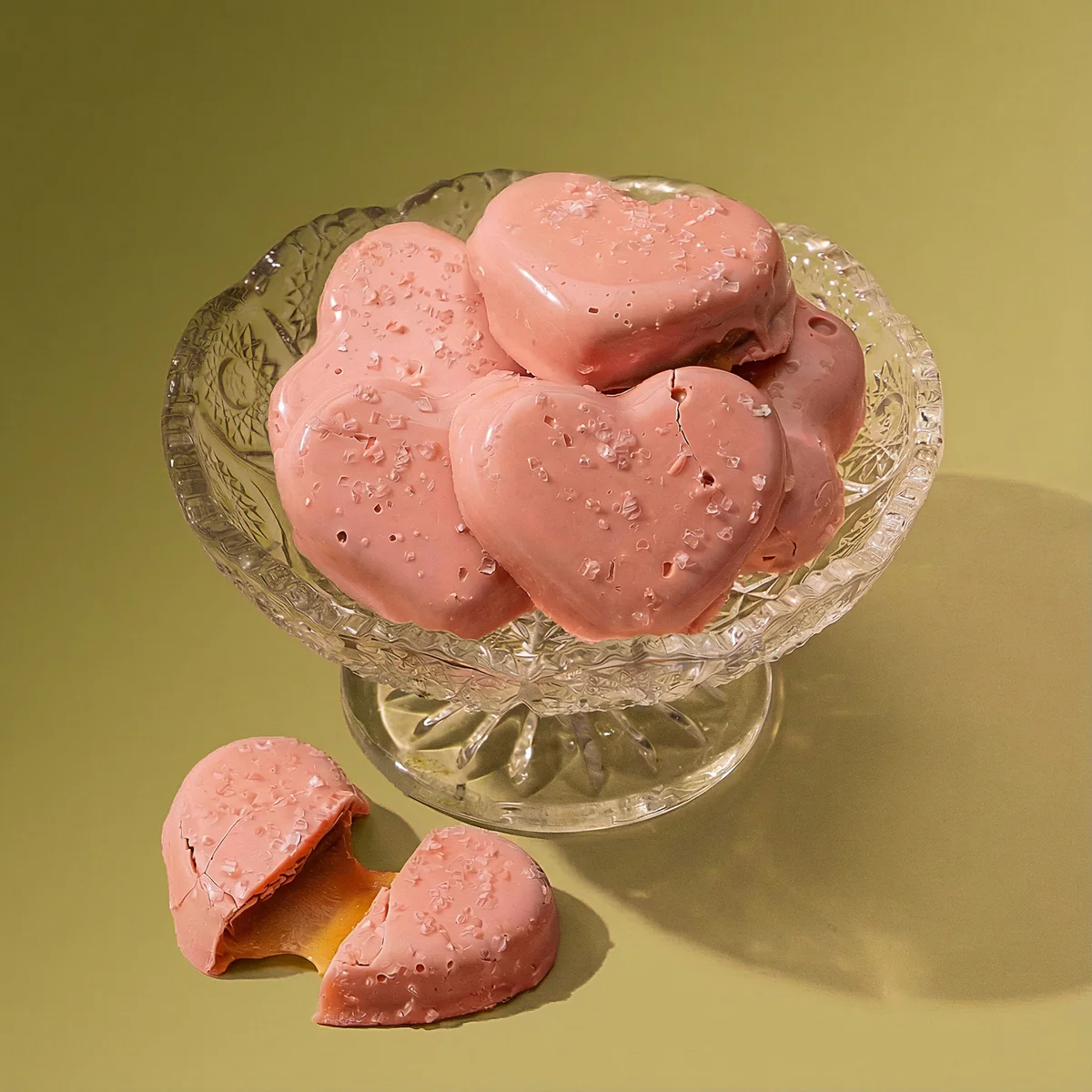 A glass dish of pink, heart-shaped chocolates with sea salt, one broken to show caramel filling.
