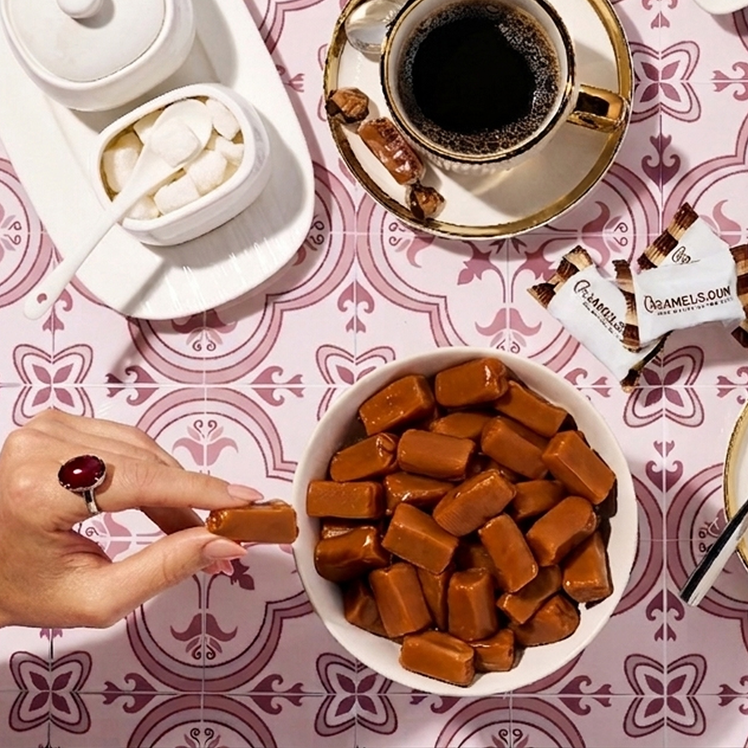 An overhead shot of a hand taking a caramel from a bowl, with a cup of coffee nearby.