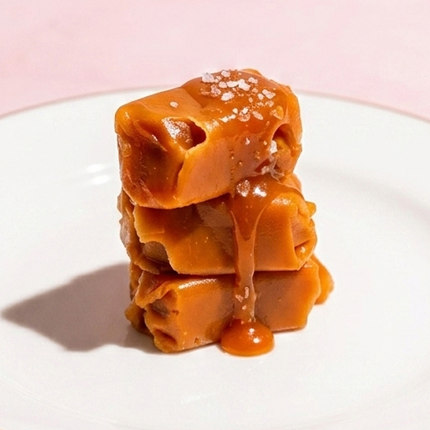 A stack of three soft caramel candies drizzled with sauce and sprinkled with sea salt on a white plate.