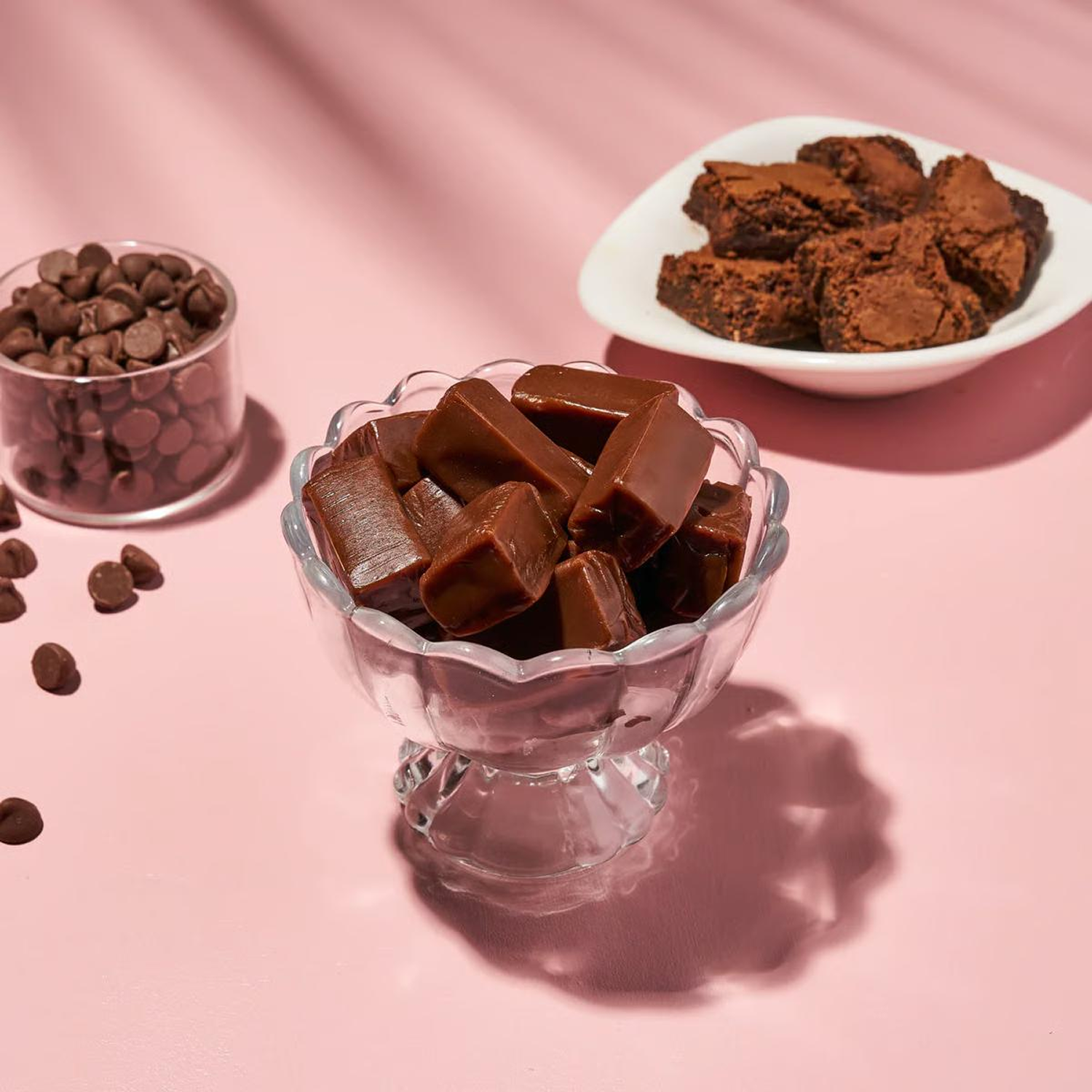 Bowls of chocolate fudge, brownies, and chocolate chips arranged on a pink surface.