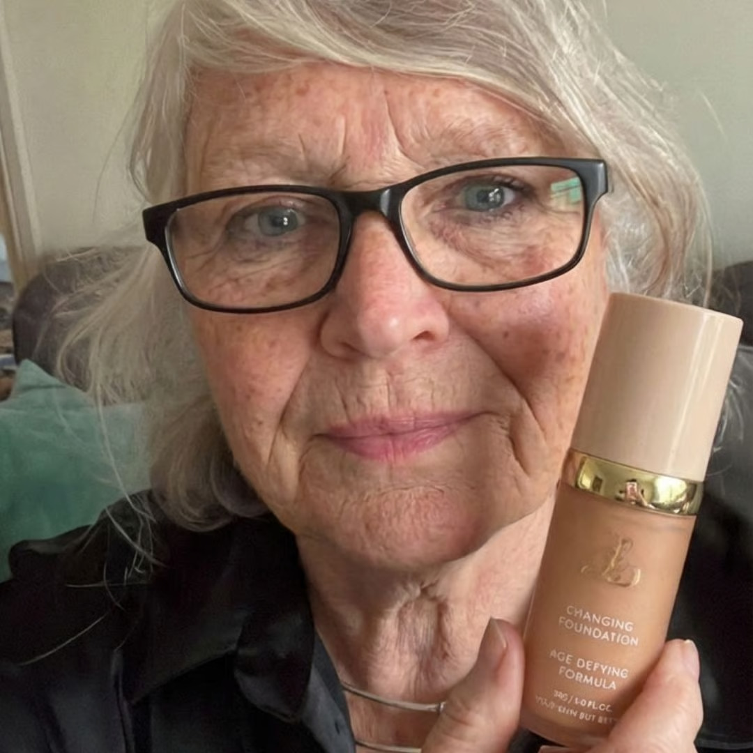 An older woman with glasses and gray hair holds up a bottle of foundation while looking at the camera.