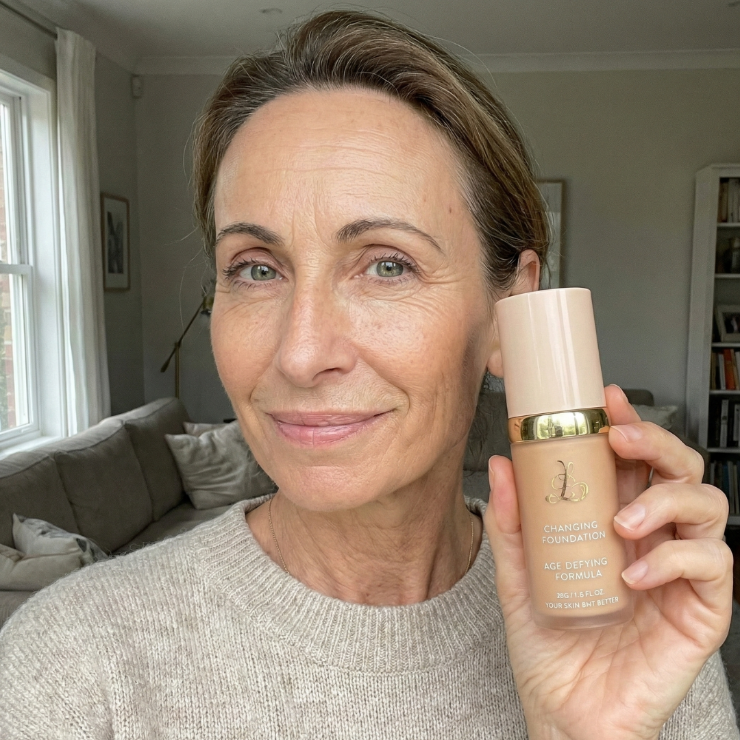 A woman with light brown hair holds a bottle of foundation while smiling in a living room.