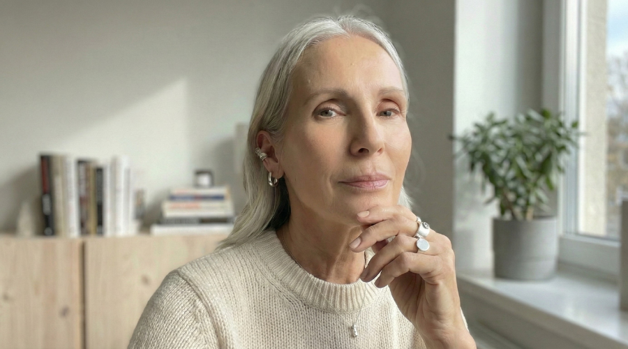 A woman with long grey hair wearing a cream sweater rests her chin on her hand, looking at the camera.