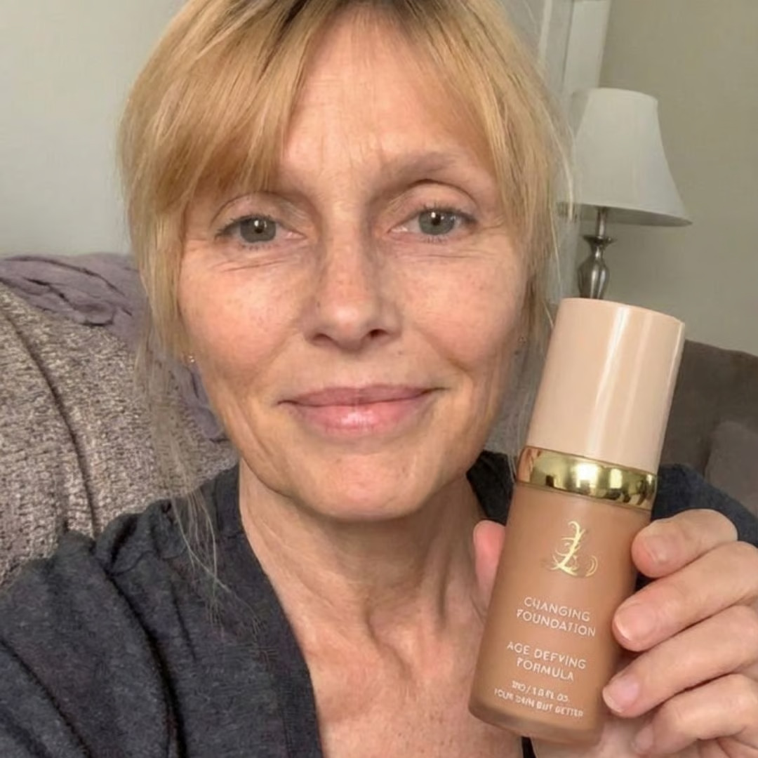 A woman with blonde hair and bangs smiles while holding up a bottle of foundation.