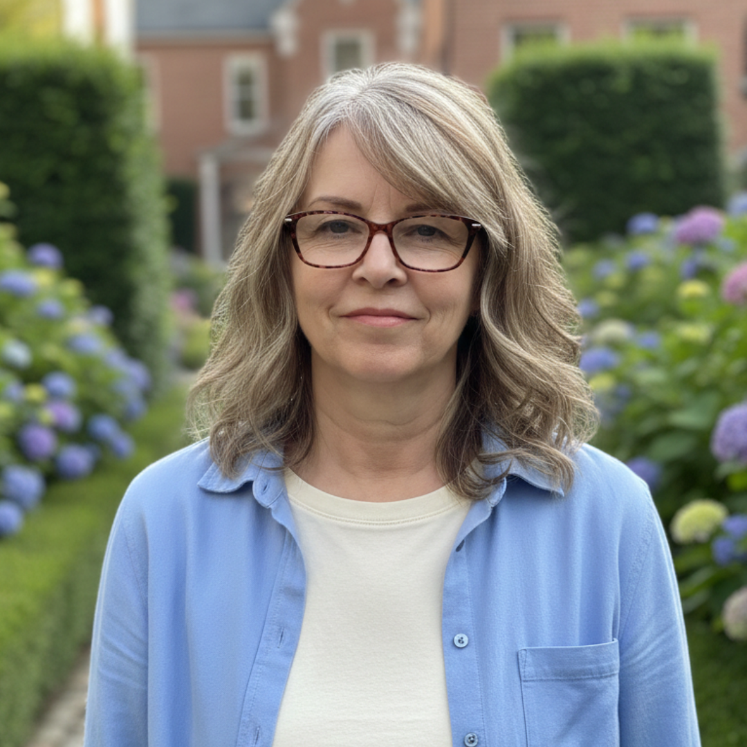 A woman with glasses and graying hair stands in a garden with blue flowers and green bushes.