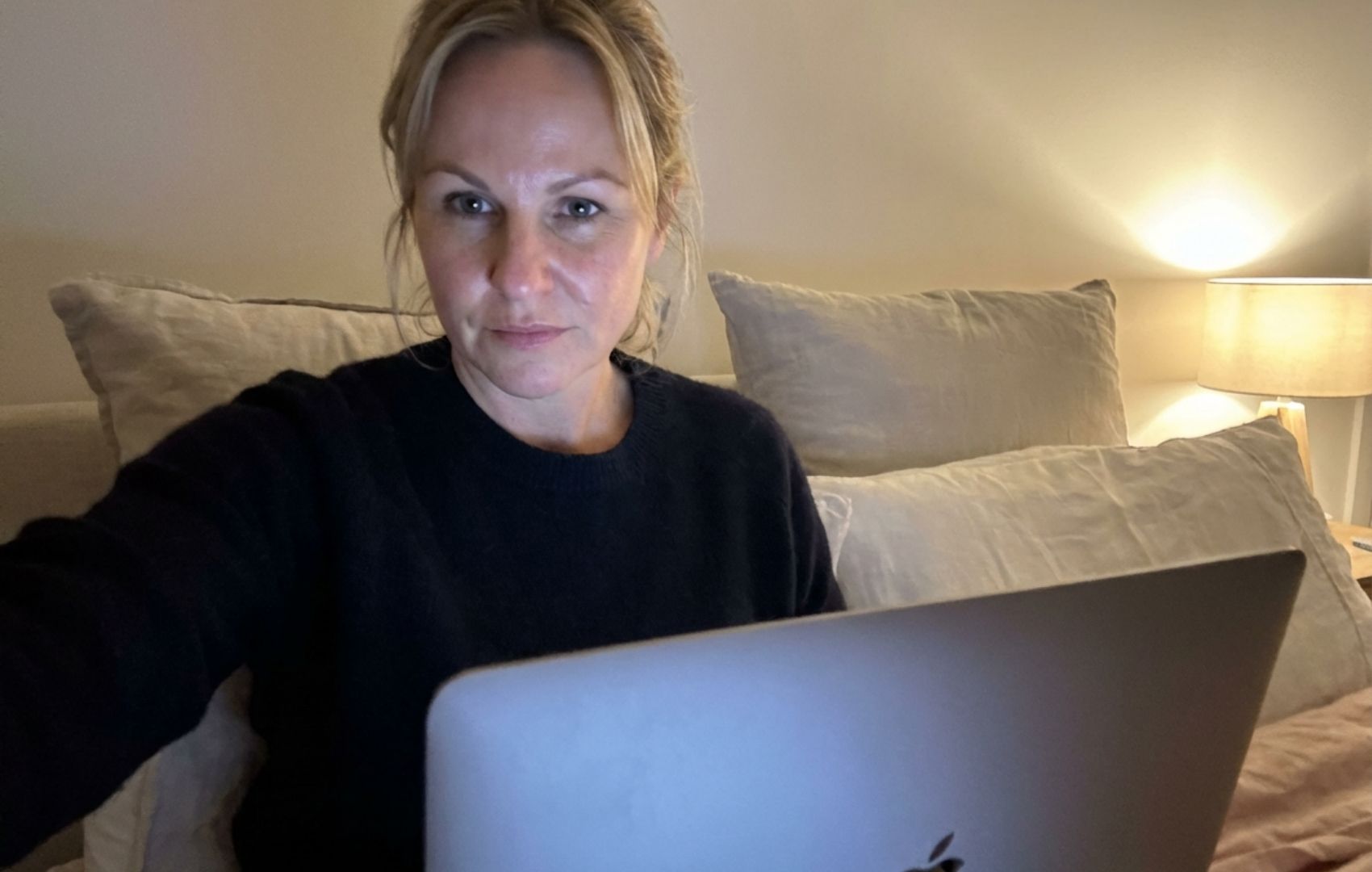 A woman in a dark sweater looks at the camera while using a laptop in a dimly lit room.