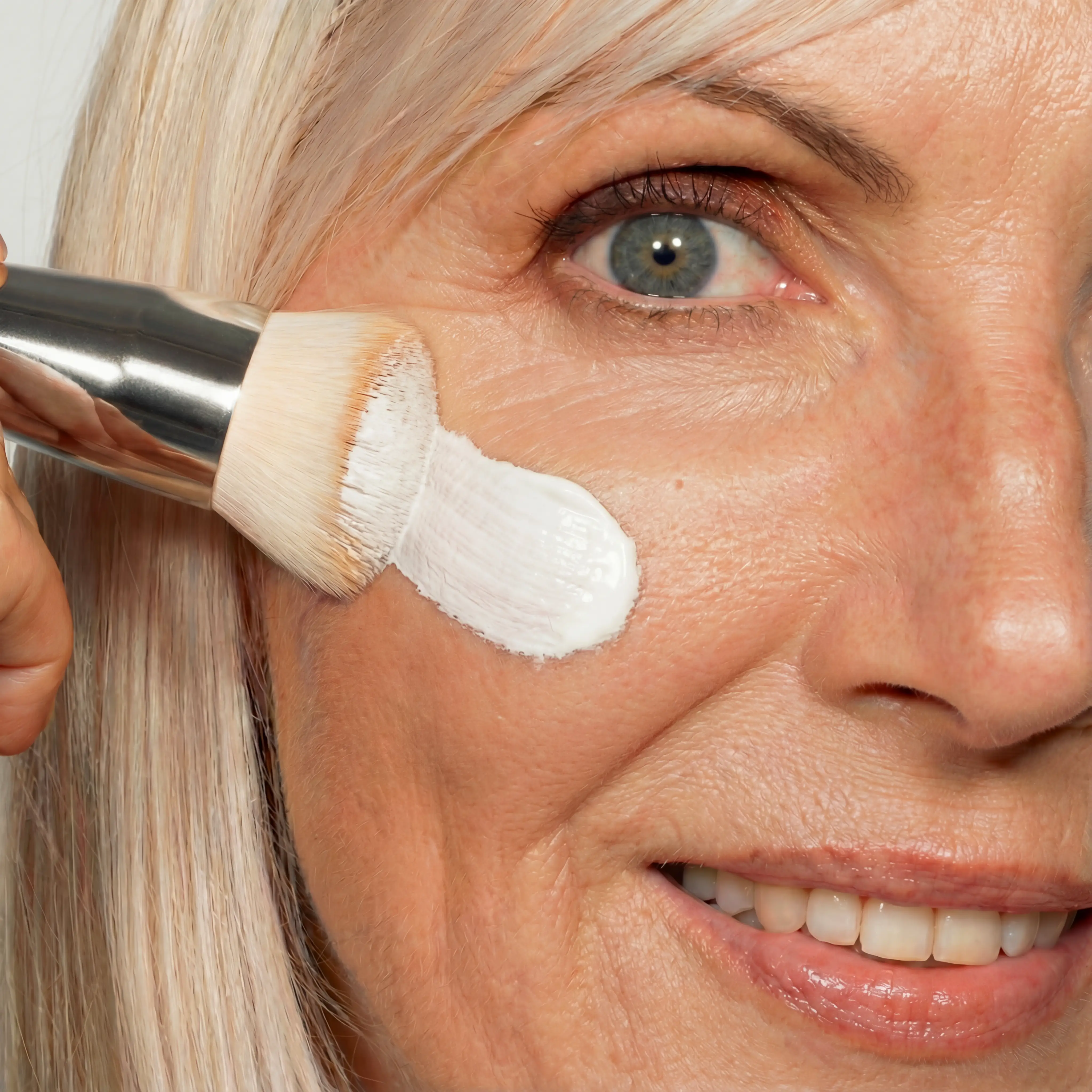 A close-up of a person applying white cream to their cheek with a brush.