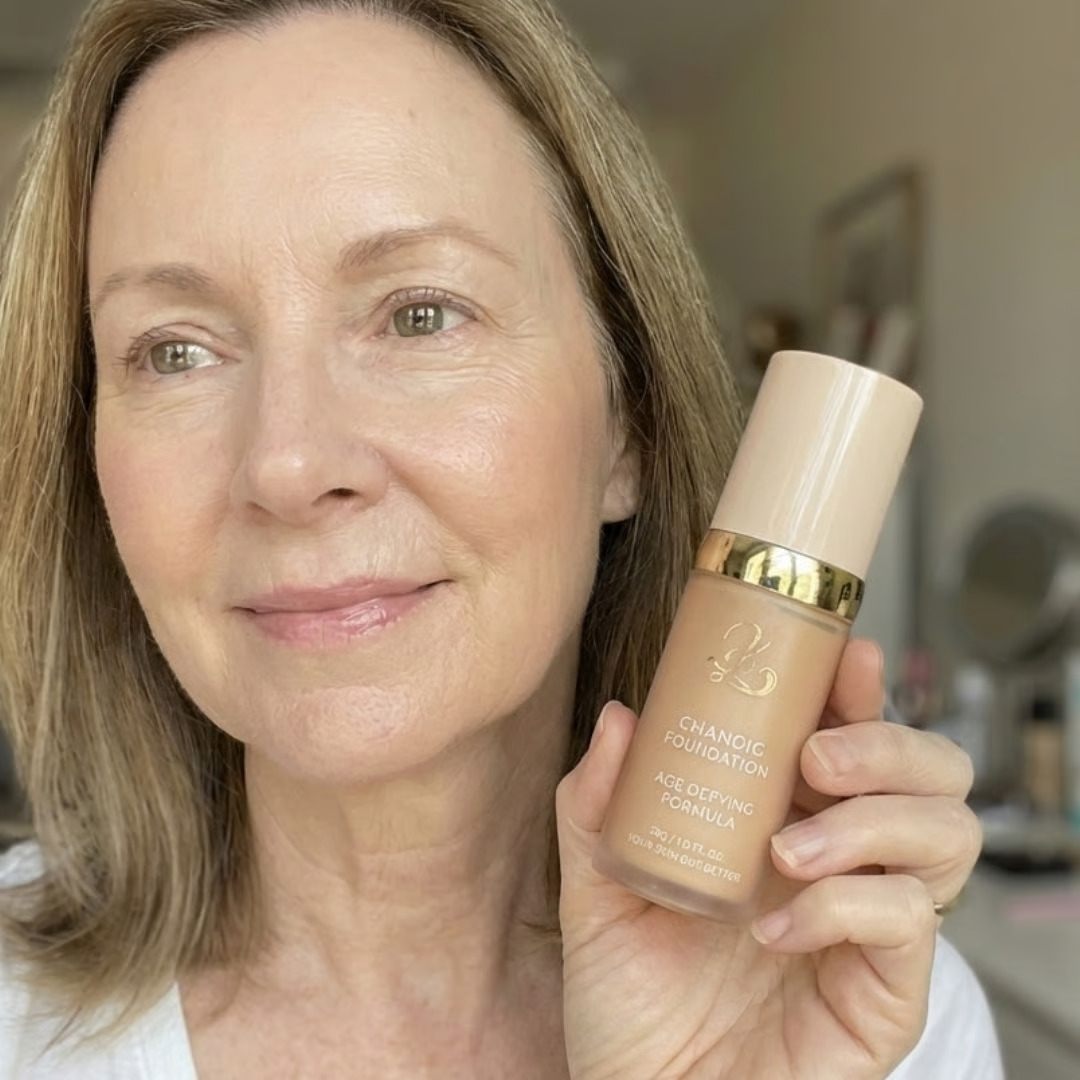 A woman with blonde hair and natural-looking makeup holds a bottle of Chanoic age-defying foundation.