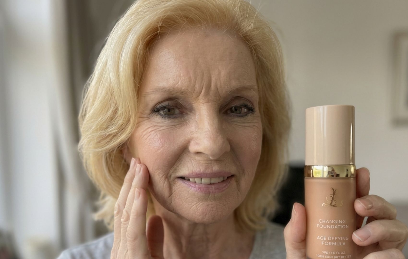 An older blonde woman smiles while holding a bottle of foundation and touching her cheek.