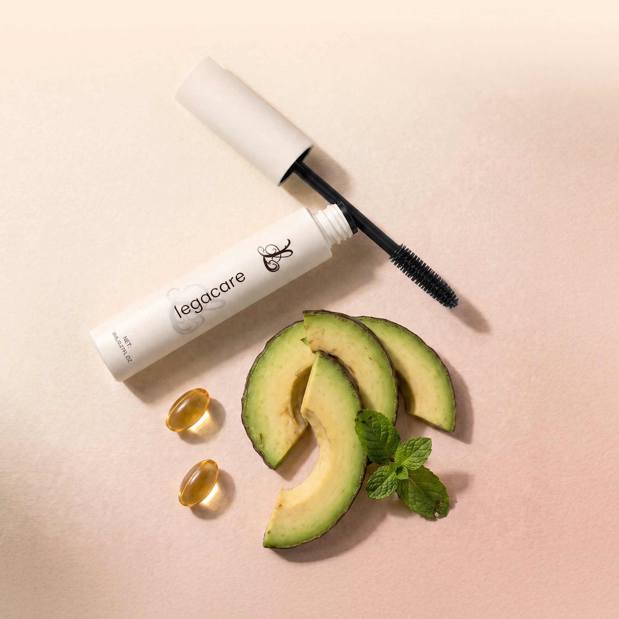 An open tube of mascara lies on a pink surface next to avocado slices, mint, and two capsules.