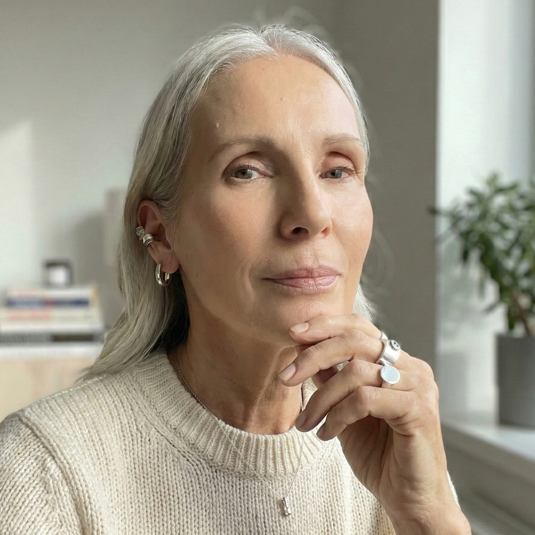 A woman with long grey hair and a cream sweater rests her chin on her hand, looking forward.