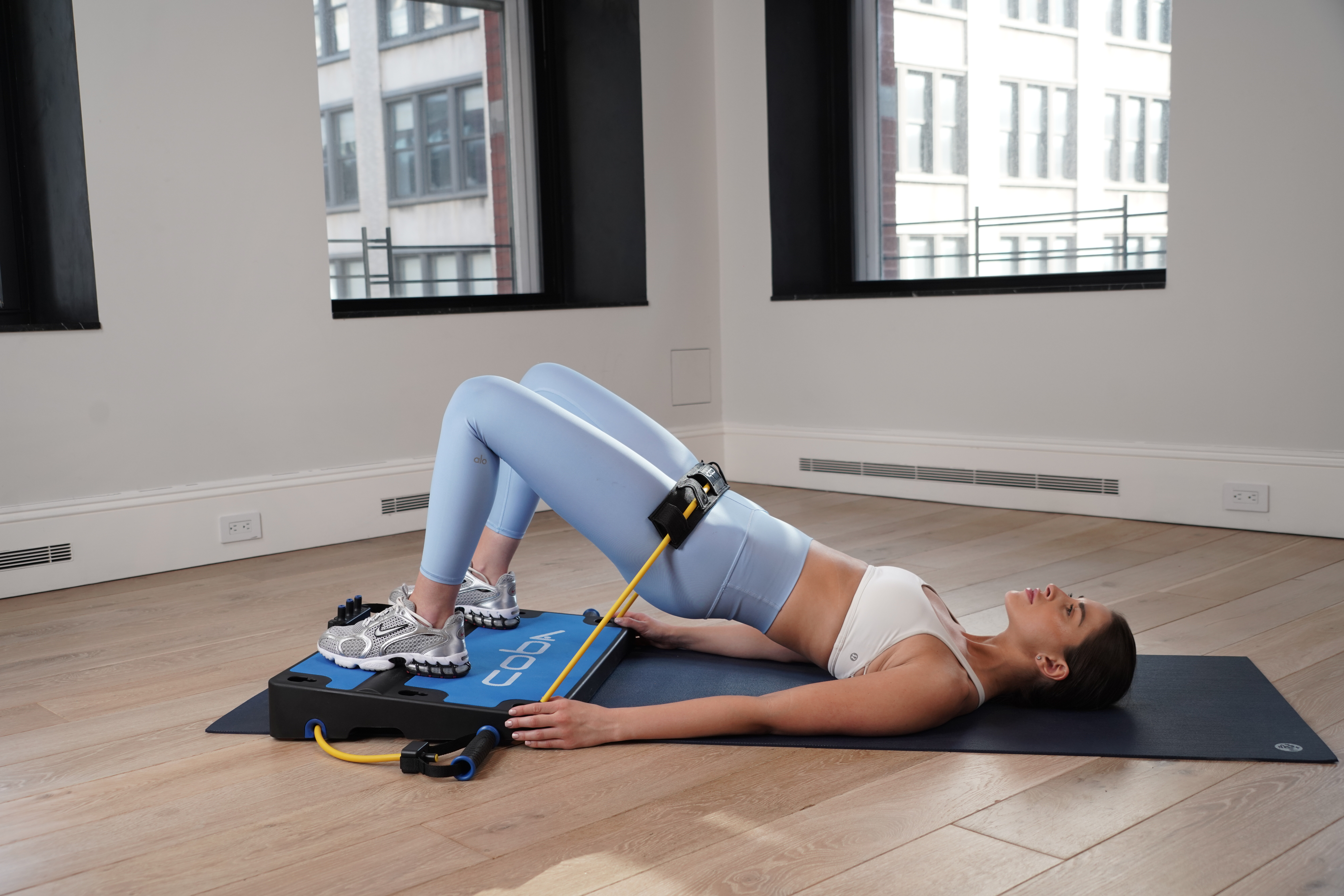 A woman performs a glute bridge on a yoga mat using a COBA board with resistance bands.