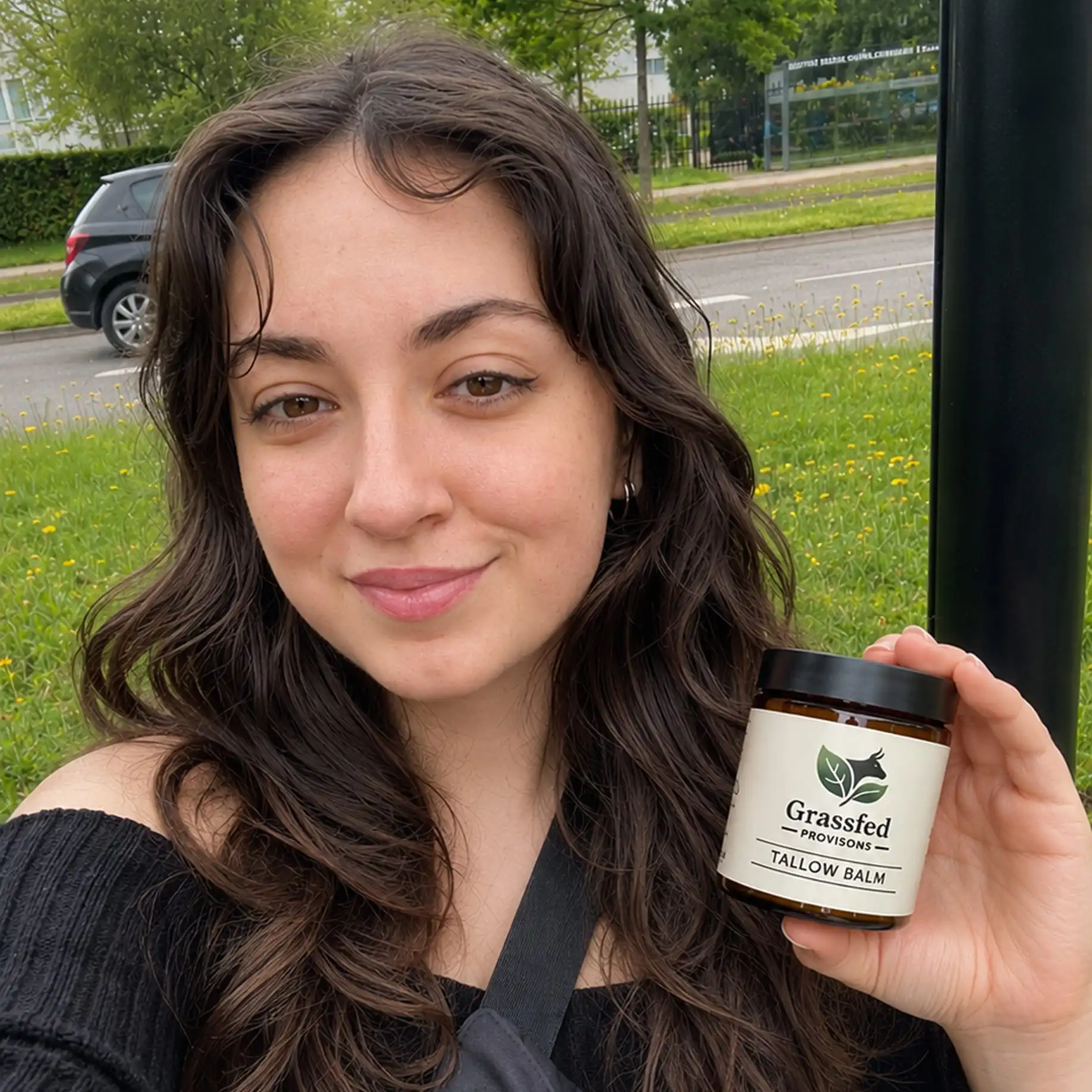 A woman with dark hair smiles and holds up a jar of Grassfed Provisions Tallow Balm.