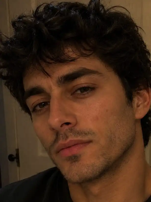 A close-up portrait of a young man with dark curly hair, brown eyes, and light facial hair.
