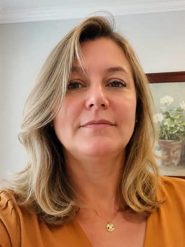 A headshot of a woman with blonde, shoulder-length hair wearing a mustard-colored top and a gold necklace.