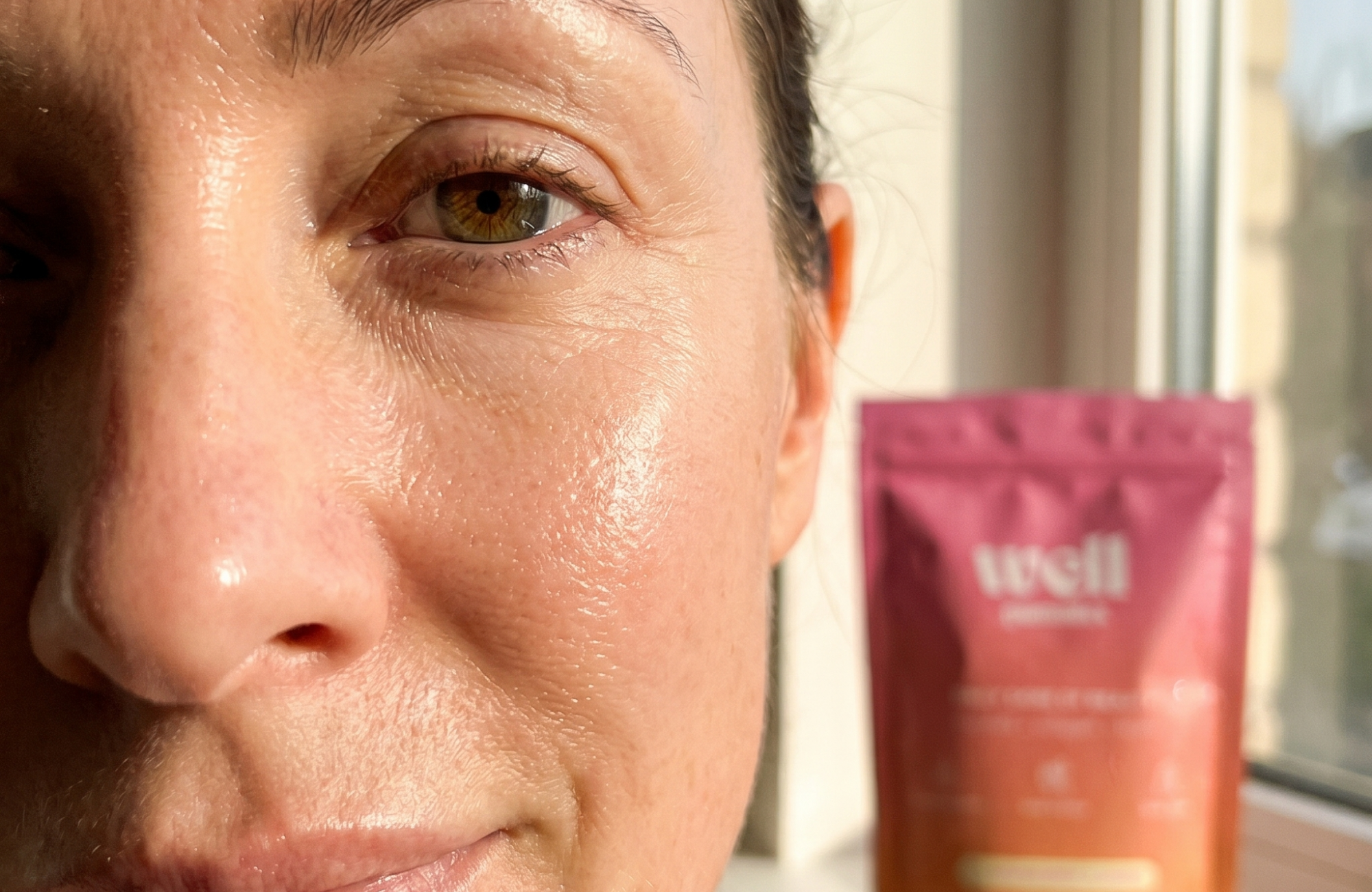 A close-up of a woman's glowing skin, with a pink product pouch blurred in the background.