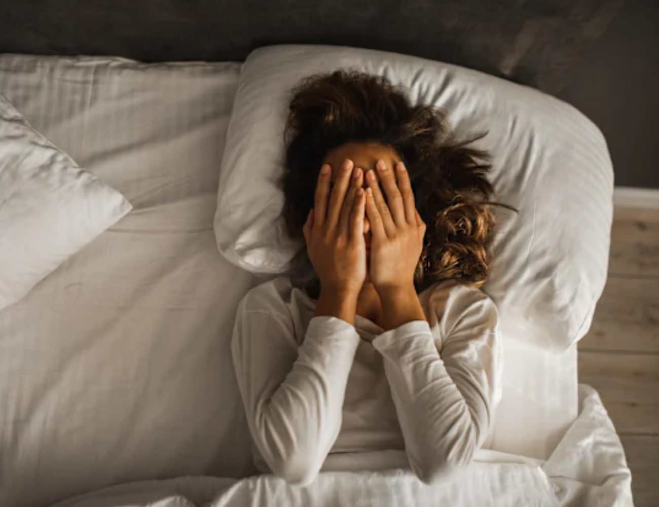 An overhead view of a person in bed covering their face with their hands.