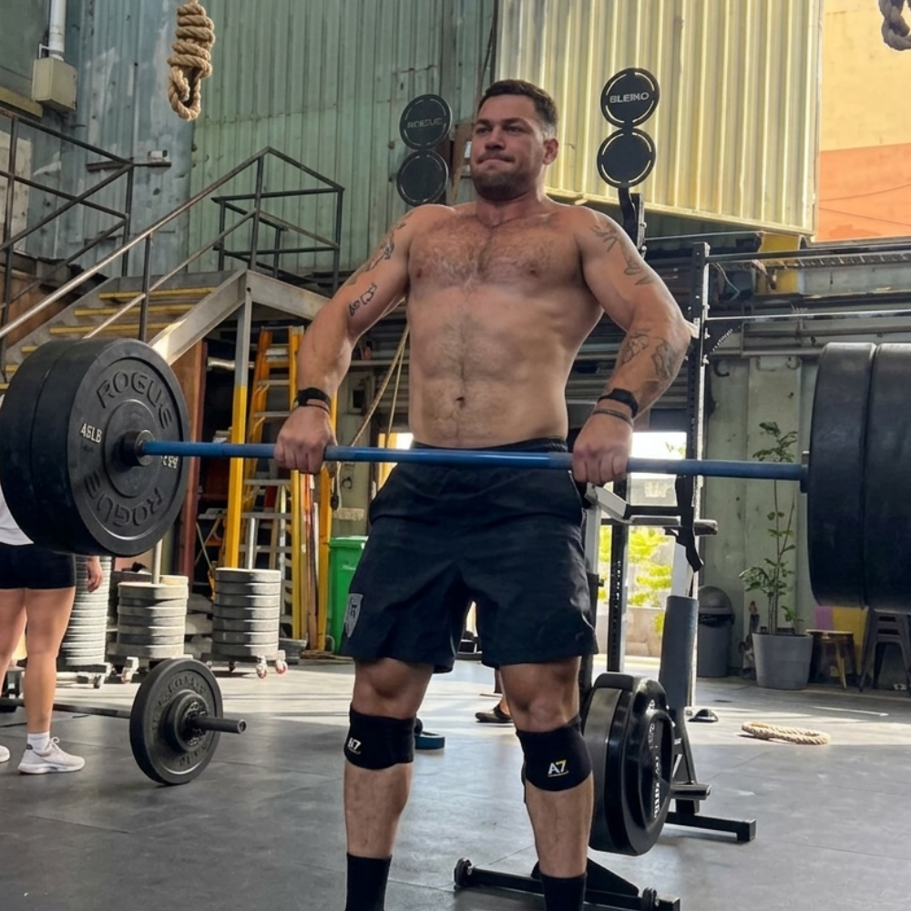 A shirtless, muscular man with tattoos lifts a heavy barbell in a gym.