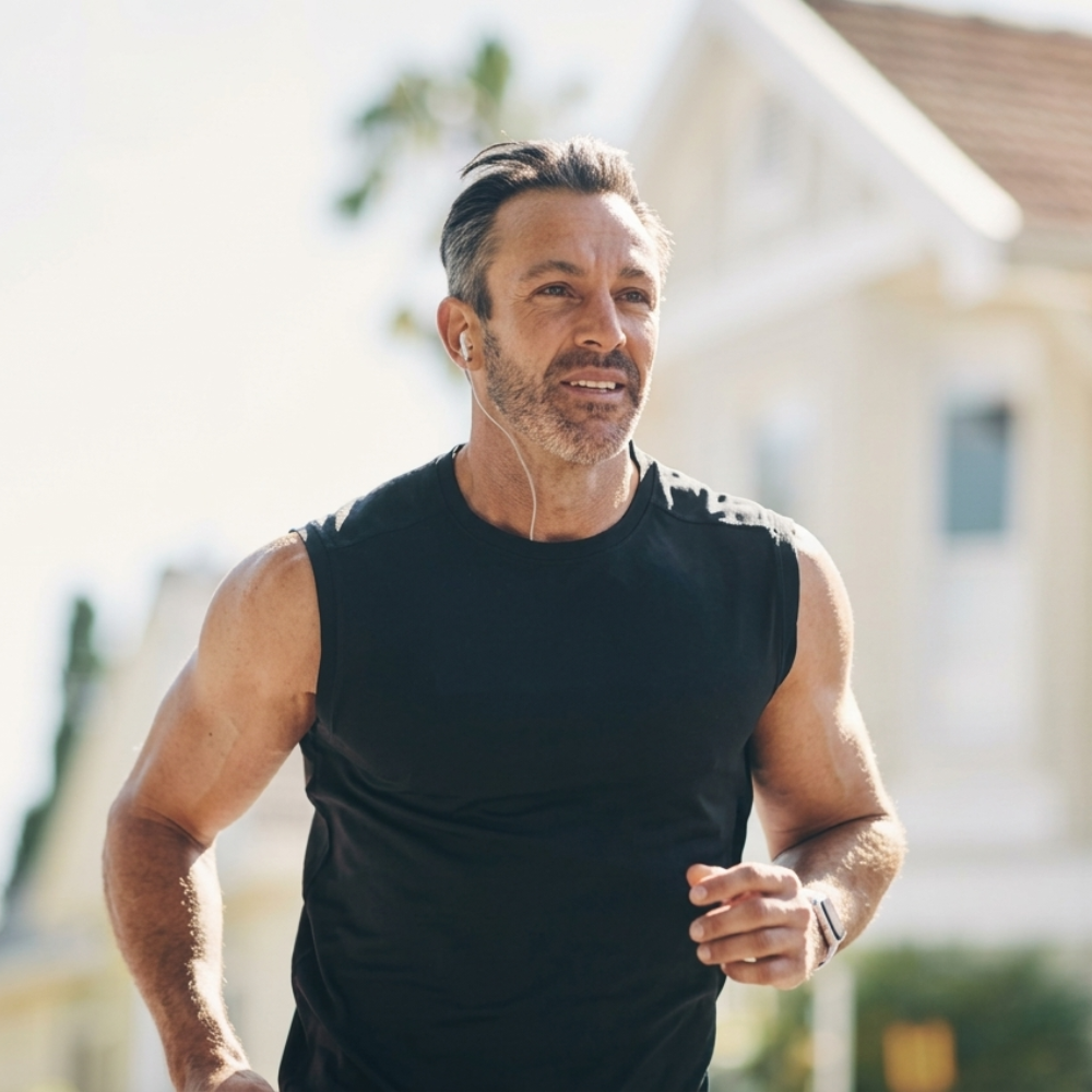 A fit, middle-aged man in a black sleeveless shirt jogs outdoors while listening to music with earbuds.
