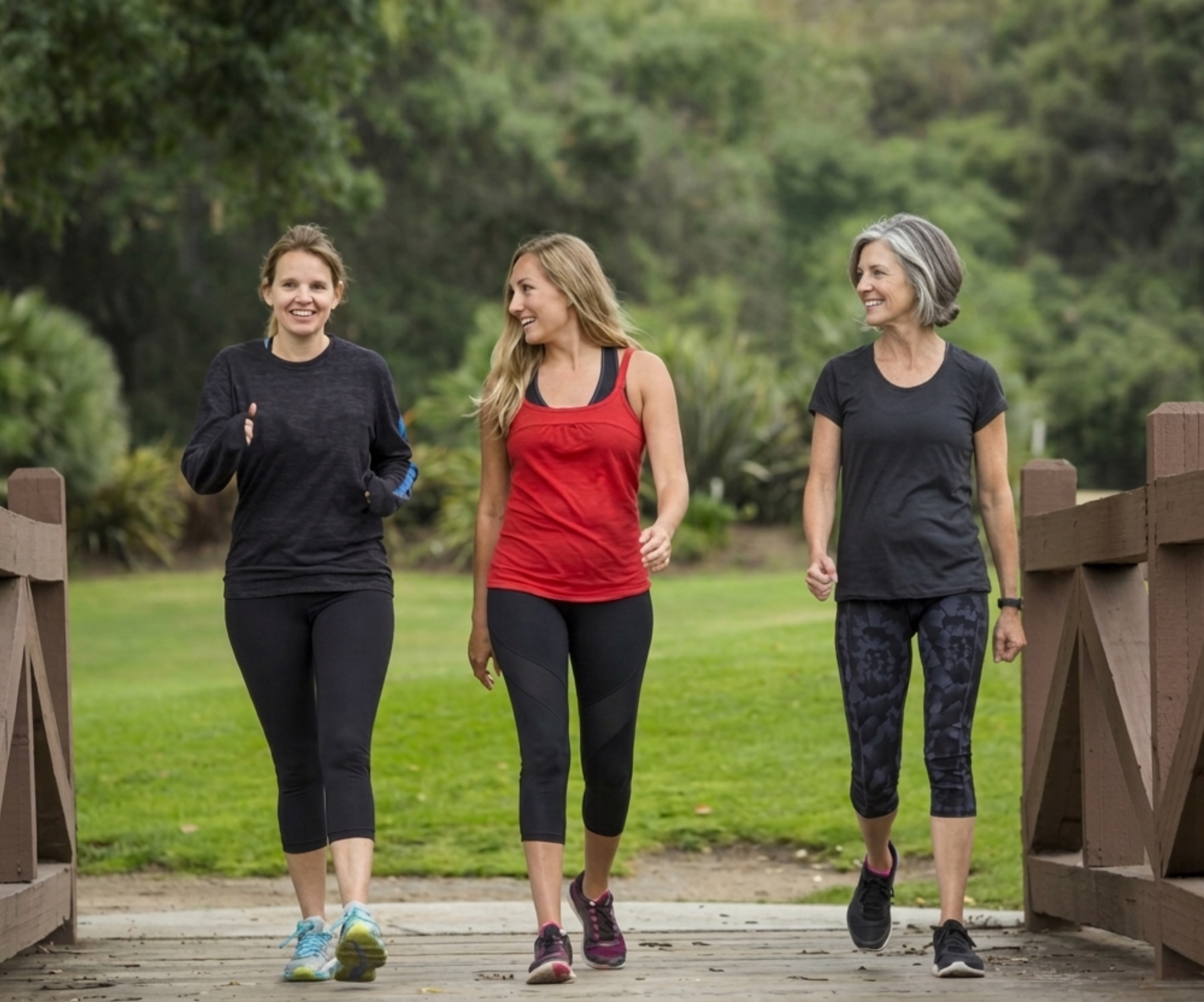 Three women in athletic wear walk together across a wooden bridge in a park.