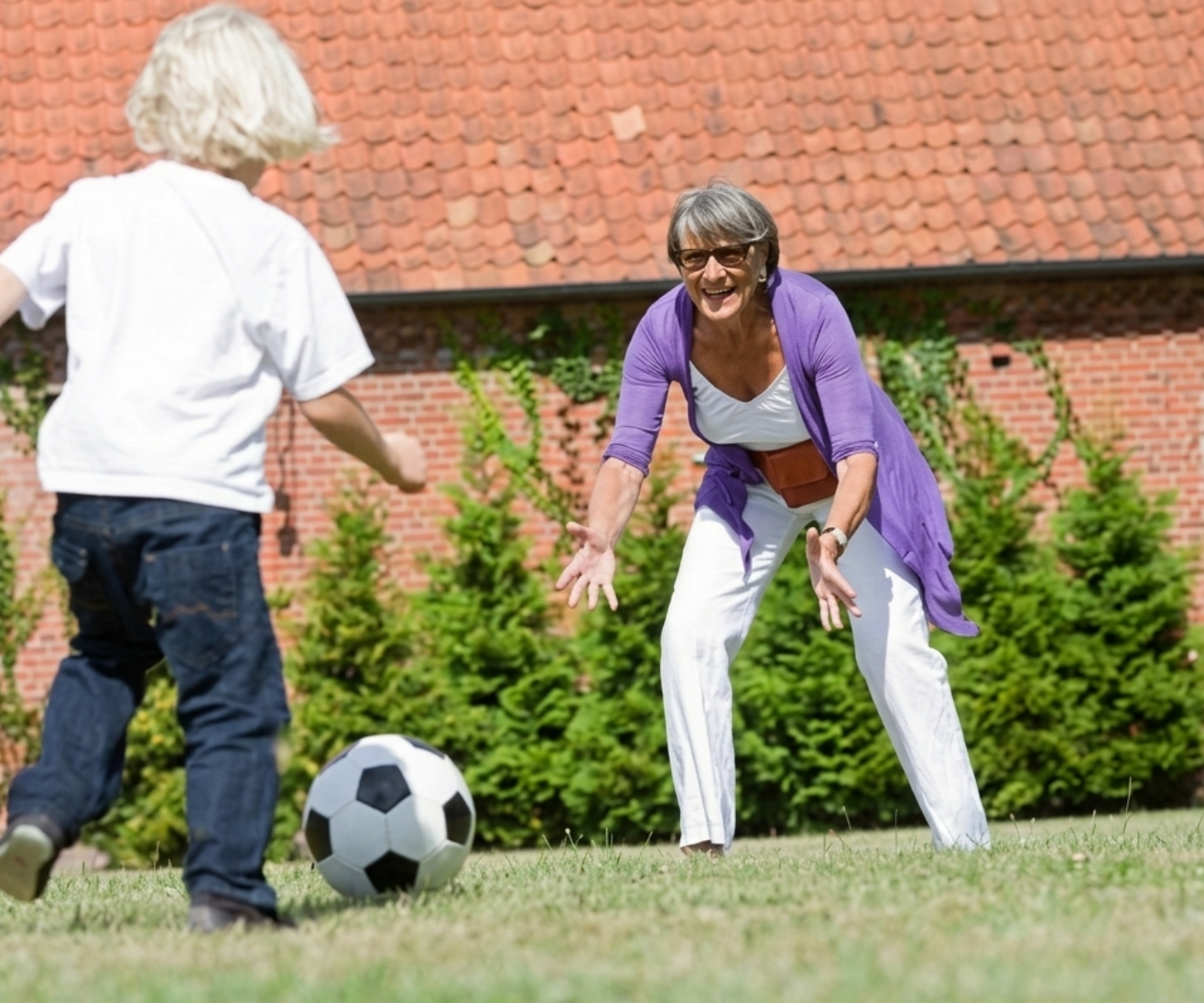 An older woman and a young child play with a soccer ball on a grassy lawn.