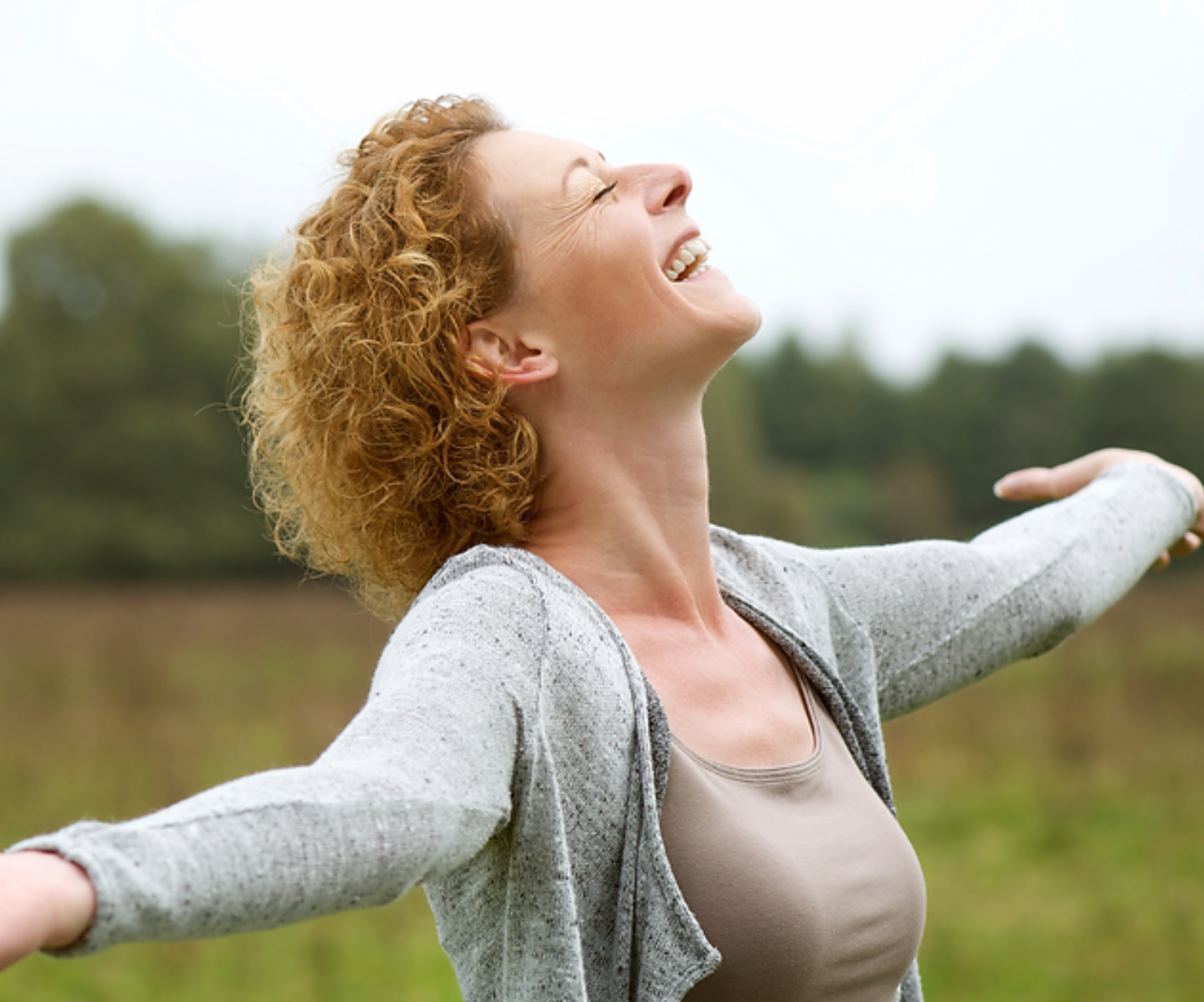 A woman with curly hair laughing with her head tilted back and arms outstretched in a field.