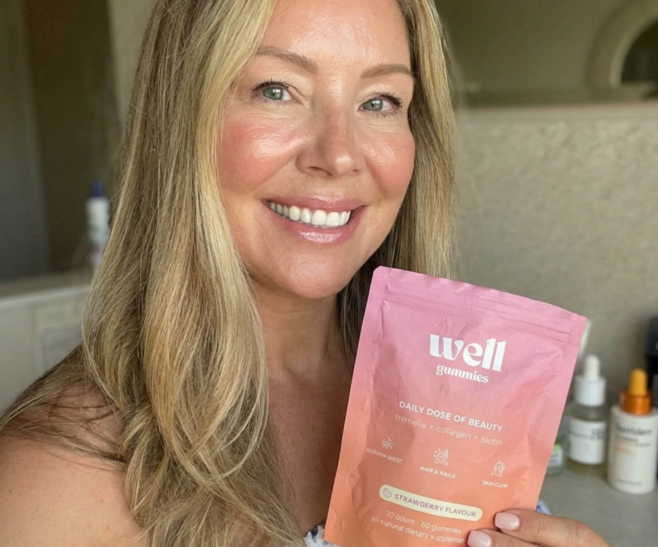 A smiling woman with blonde hair holds a pink pouch of Well Gummies dietary supplements.