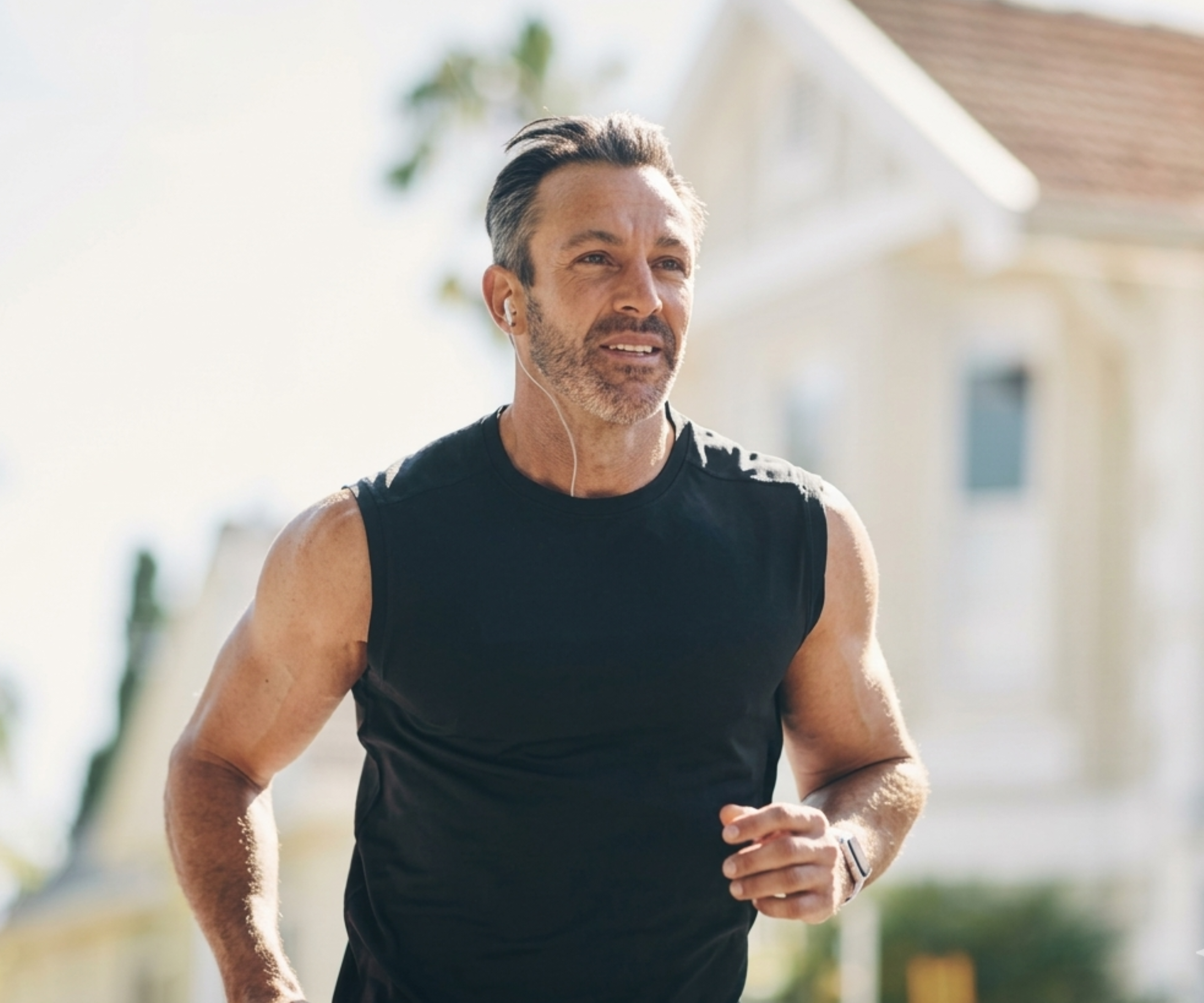 A middle-aged man in a black sleeveless shirt jogs outdoors while listening to earbuds.